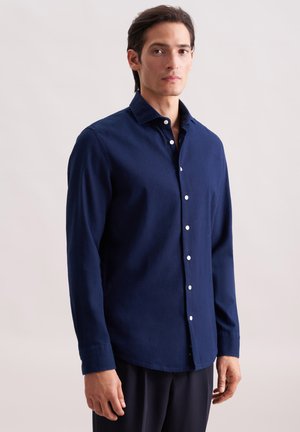 Un uomo in camicia a maniche lunghe blu navy con bottoni e pantaloni neri, in piedi e rivolto in avanti, su uno sfondo chiaro e semplice.