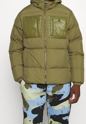 Doudoune vert olive avec poches poitrine doubles brillantes, fermeture éclair frontale et capuche, portée avec un pantalon camouflage bleu, noir et vert.