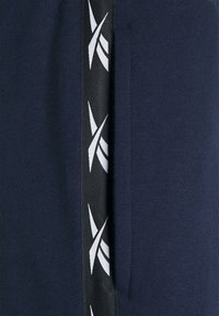 Námořnické modré tepláky s boční kapsou a černým pruhem s bílými grafickými akcenty na boku. Měkká, texturovaná tkanina.