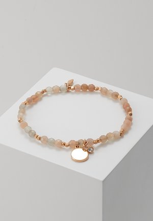 Bracelet avec des petites perles beiges et dorées alternées, agrémenté d'un disque rond en or et d'un petit pendentif en cristal, présenté sur un bloc blanc.
