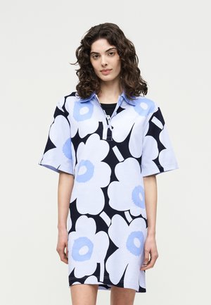 Mujer con cabello rizado que lleva un vestido de manga corta hasta la rodilla con un estampado floral grande en azul y blanco sobre un fondo negro.