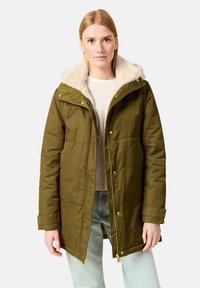 Parka de color verde oliva con capucha forrada de piel sintética, con cierre de cremallera, botones a presión y bolsillos laterales, llevada sobre un suéter ligero.