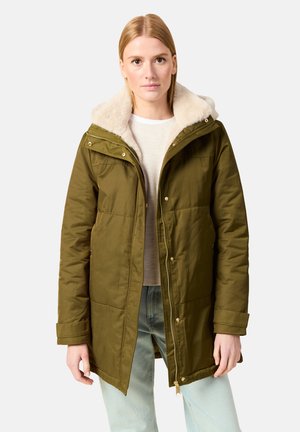 Olijfgroene parka met een nepbont gevoerde capuchon, met een rits aan de voorkant, drukknopen en zijzakken, gedragen over een lichte trui.
