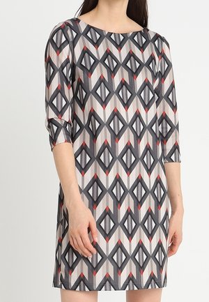 Kvinde iført en knælængde kjole med trekvartærmer og et geometrisk diamantmønster i sort, grå, hvid og små røde accenter.