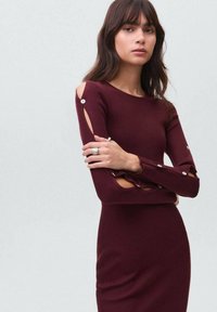 Rochie burgundy cu model nervurat și mâneci lungi, având un design cu umeri decupați și accente cu nasturi. Material moale, texturat, cu o siluetă ajustată.