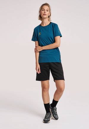 Tyrkis kortærmet træningsshirt med sorte detaljer, parret med sorte shorts. Har logo detaljer på shirtet. Modellen bærer sporty sneakers og strømper.
