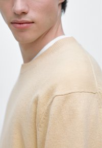 Béžový pletený svetr s kulatým výstřihem, s hladkou texturou a jemnými šicími detaily. Minimalistický design, pohled zblízka.