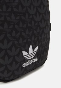 Mochila de texto negra con una superficie estampada con un diseño floral repetido. Incluye un logo de Adidas en blanco en la parte inferior.