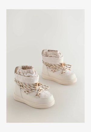 Fehér bokacsizmák tűzött textúrával, rugalmas szegéllyel és arany díszítésű fűzőkkel. Kerek orrúak és gumitalppal rendelkeznek a tapadás érdekében.