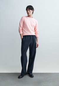 Mlajša oseba, oblečena v roza majico z dolgimi rokavi, črne hlače in črne čevlje, stoji z eno roko v žepu pred belim ozadjem.