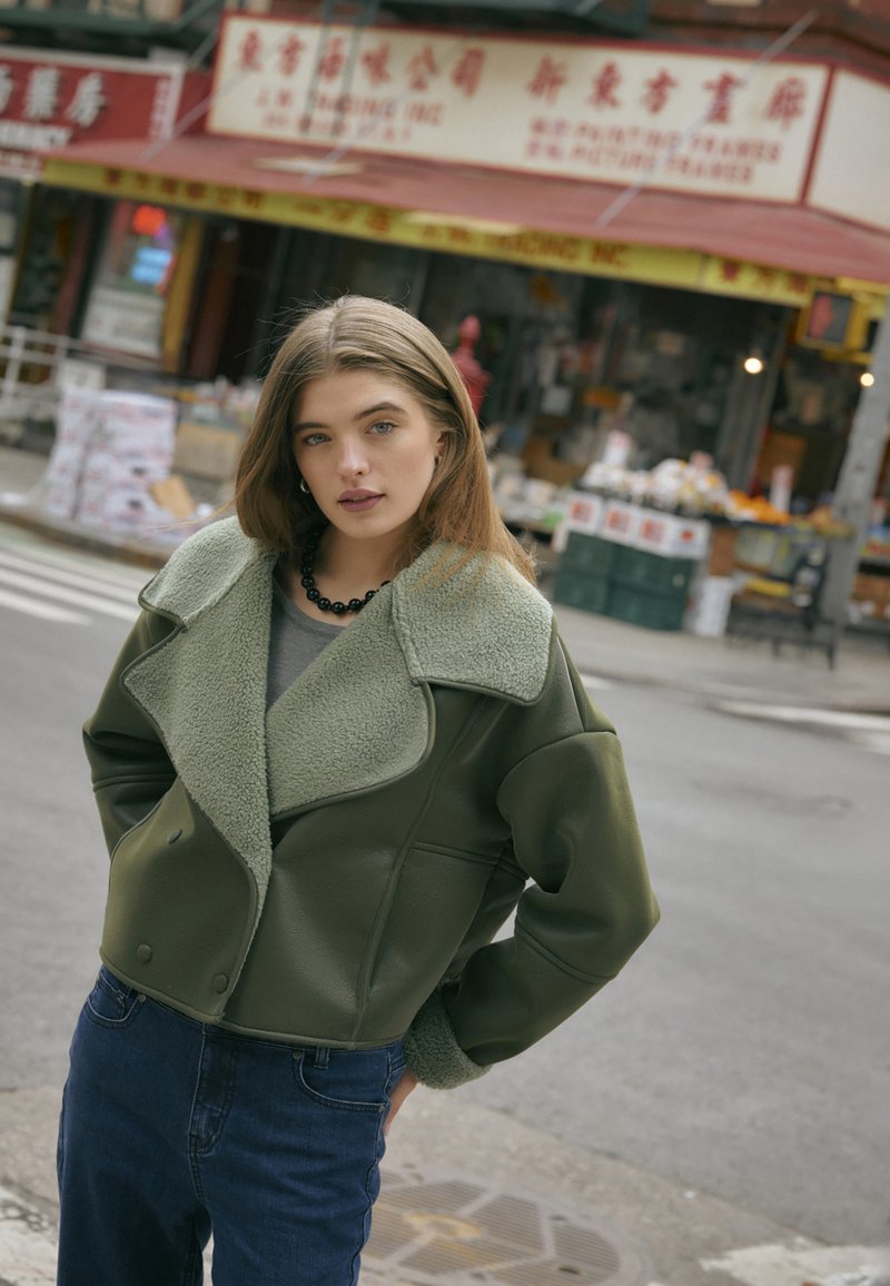 Oliwkowa krótka kurtka z miękkim, teksturowanym jasnozielonym kołnierzem. Połączona z wysokim stanem ciemnych jeansów i czarnym naszyjnikiem z koralików.