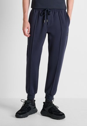 Man draagt marineblauwe joggingsbroek met elastische taille en zwarte sportschoenen, staand tegen een effen achtergrond.