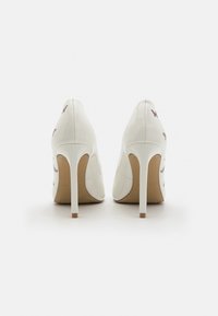 Escarpins en cuir verni blanc avec des pointes effilées, des talons aiguilles et des accents décoratifs en forme de papillon sur les côtés.