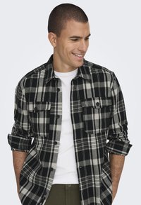 Cămașă flanelată în carouri alb-negru, cu două buzunare pe piept, mâneci rulote și închidere cu butoane. Purtați-o peste un tricou alb.