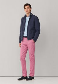 Veste zippée marine, pull bleu clair, chinos roses et chaussures noires sans lacets. Design minimaliste avec une coupe décontractée et des textures lisses.
