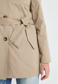 Beżowy trencz z paskiem w talii, z dużymi guzikami i gładką fakturą, zaprezentowany na dżinsach.