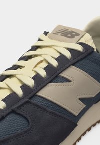 Gros plan d'une basket New Balance avec des lacets beiges, un tissu en mesh bleu marine, des empiècements en daim foncé et un grand logo beige "N" sur le côté.