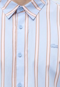 Chemise bleu clair à col avec des rayures verticales roses et marron, des boutons bleus et un petit logo crocodile brodé sur la poitrine.