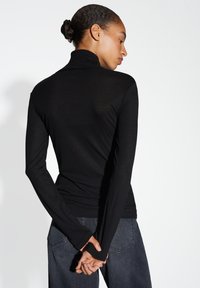 Femme avec les cheveux en chignon portant un haut à col roulé noir à manches longues et un jean foncé, se tenant les mains jointes derrière le dos, tournée vers l'arrière.
