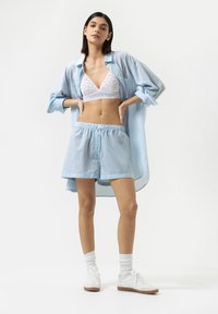 Lichtblauw gestreepte oversized blouse, witte kanten bralette, bijpassende lichtblauwe shorts en witte sneakers met bruine zolen. Diagonale perspectief.