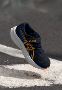 Ναυτικό μπλε αθλητικό παπούτσι με διχτυωτό άνω μέρος, καμπυλωτή λευκή σόλα, κίτρινες λεπτομέρειες και ιμάντα με Velcro, να αιωρείται πάνω από μια υφασμάτινη επιφάνεια.