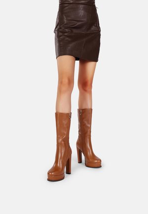Stivali marroni in pelle al ginocchio con tacco alto, caratterizzati da una cerniera laterale e una texture liscia. Abbinati a una minigonna in pelle marrone.