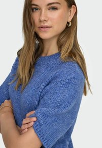 Modrý svetr z texturované pleteniny s krátkými rukávy a kulatým výstřihem, doplněný zlatými kruhovými náušnicemi.