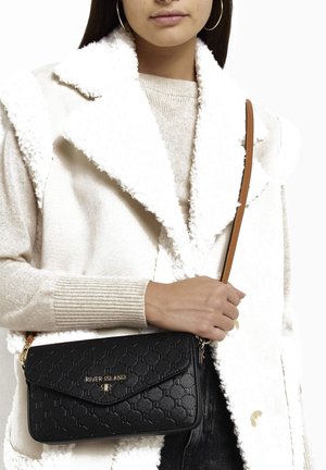 Svart texturerad axelremsväska med guldfärgad metall och logotyp, med en klaffstängning. Bärs med en vit texturerad kappa och beige tröja.