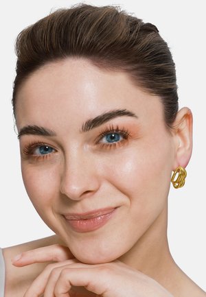 Goldene Creolen mit verdrehtem Design, getragen von einer Person mit heller Haut und dunklem Haar. Glatte Textur und glänzende Oberfläche.