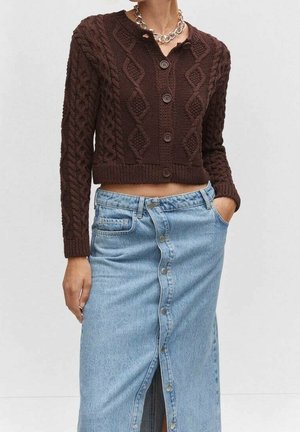 Femme portant un cardigan brun à boutons en maille torsadée et une jupe taille haute en denim bleu clair à boutons sur le devant, les mains dans les poches.