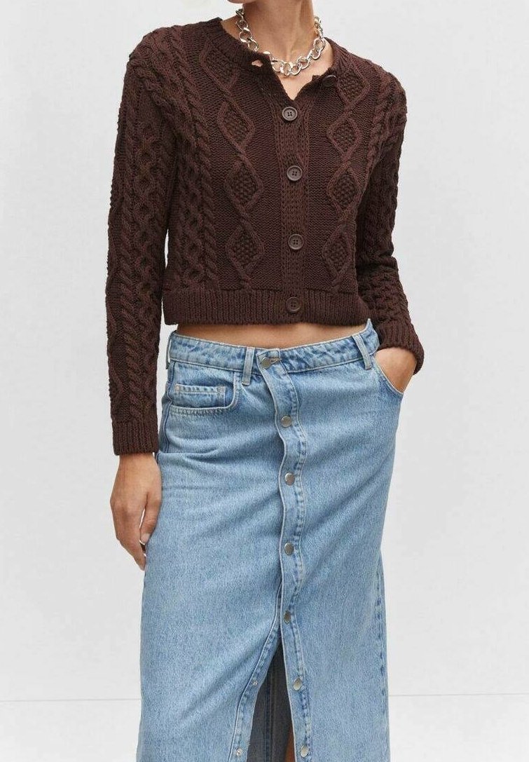 Femme portant un cardigan brun à boutons en maille torsadée et une jupe taille haute en denim bleu clair à boutons sur le devant, les mains dans les poches.