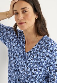Blauwe bloemenblouse met een v-hals, voorzien van kleine witte en zwarte patronen op een lichtblauwe achtergrond. Zachte, lichte stof.