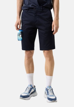 Man draagt marineblauwe korte broek, witte enkelsokken en blauw-witte sneakers, houdt reflecterende blauwe zonnebril in linkerhand.