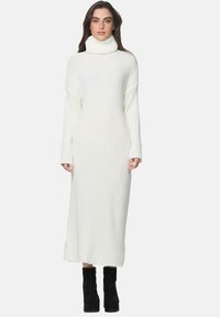 Robe à col roulé côtelée blanche, à manches longues, avec des fentes sur les côtés, longueur cheville, texture douce, associée à des bottes compensées noires.