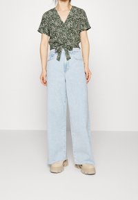 Top avvolgente floreale verde con maniche corte e un nodo in vita, abbinato a jeans a gamba larga blu chiaro e scarpe chunky beige.