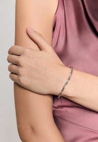 Srebrna bransoletka o skręconym wzorze, z delikatnymi ogniwami i zapięciem typu lobster, noszona na ramieniu o gładkiej, jasnej karnacji.