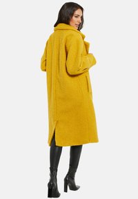 Abrigo de forro polar amarillo con una superficie texturizada, cuello amplio, mangas largas y bolsillos laterales; cuenta con una abertura trasera para mayor movilidad.