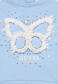 Ανοιχτό μπλε φούτερ με λευκό σχέδιο πεταλούδας με βολάν και πούλιες και το κείμενο "GUESS" σε μεταλλικό φύλλο παρακάτω.