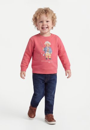 Tout-petit souriant aux cheveux bouclés portant un sweat corail avec un motif ours, un jean foncé et des baskets marron, marchant vers l'avant sur un fond blanc.