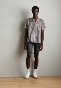 Camisa de manga corta con estampado en blanco con puntos oscuros, combinada con pantalones cortos de mezclilla oscuros. Zapatillas blancas con suelas texturizadas completan el atuendo.