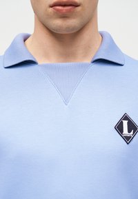 Homme portant une chemise bleu clair à col avec une insertion en V côtelée et un écusson noir en forme de losange du logo Lacoste sur la poitrine.