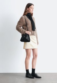 Brauner, flauschiger Cardigan, schwarzer Schal, cremefarbener Leder-Minirock, schwarze Schnürschuhe und schwarze Kniestrümpfe; minimalistischer Stil.