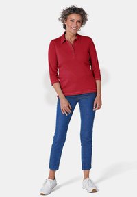 Rode polo-shirt met driekwartmouwen, een klassieke kraag en knoopsluiting, gecombineerd met slanke blauwe spijkerbroeken en witte sneakers.