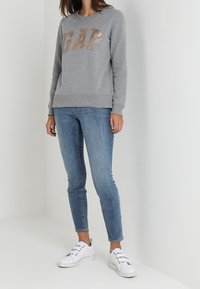 Mujer con un sudadera gris de GAP, jeans ajustados azules y zapatillas blancas, de pie contra una pared blanca.