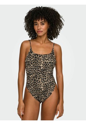 Donna con capelli ricci che indossa un costume intero con stampa leopardata e sottili spalline, in piedi davanti a uno sfondo chiaro e uniforme.