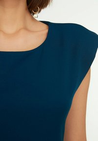 Tyrkysové šaty s širokým, zaobleným výstřihem a strukturovanými krátkými rukávy. Hladká tkanina a jemný lesk, doplněné visícími náušnicemi s korálky.