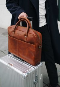 Brown leather briefcase with double handles, zipper closure, and front pocket, resting on a silver metallic suitcase. Smooth texture.