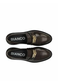 Bruine leren instappers met een gouden kettingaccent en franjes aan de voorkant, met een zwarte binnenzool waarop de merknaam "BIANCO" is weergegeven.