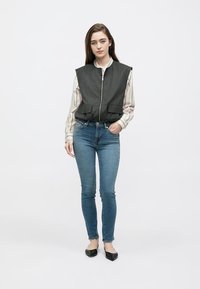 Siva brezrokavnik z zadrgo, širokimi rameni in sprednjimi žepi, nošen preko svetle srajce z črtami. Kombiniran z oprijetimi modrimi jeans hlačami in črnimi čevlji brez pete.