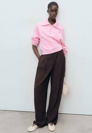 Maglione rosa a zip, pantaloni larghi marroni e scarpe chiare. L'outfit presenta linee rilassate e un piccolo borsa intrecciata come accent.